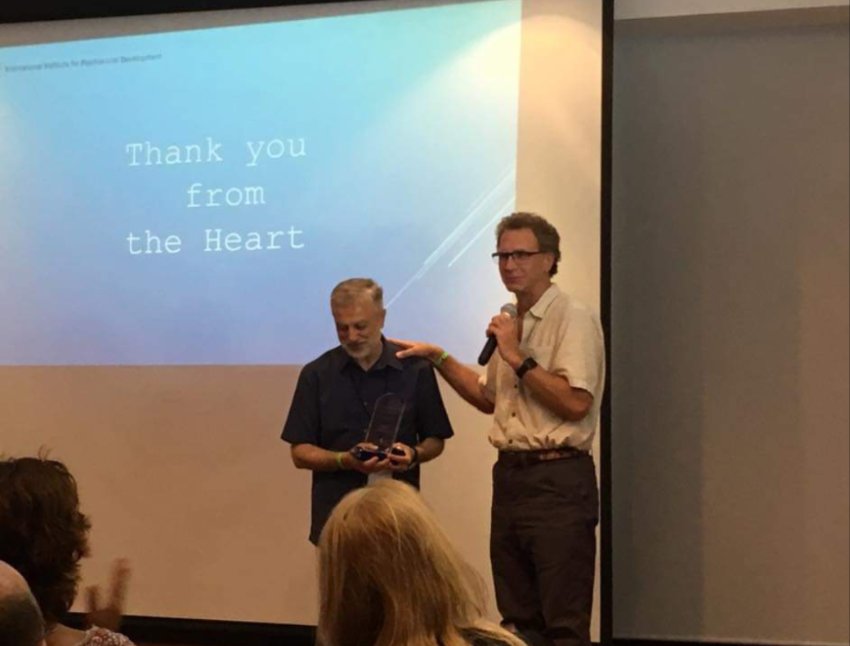 Majd on stage beside a speaker, holding an award, beneath a screen reading Thank you from the Heart